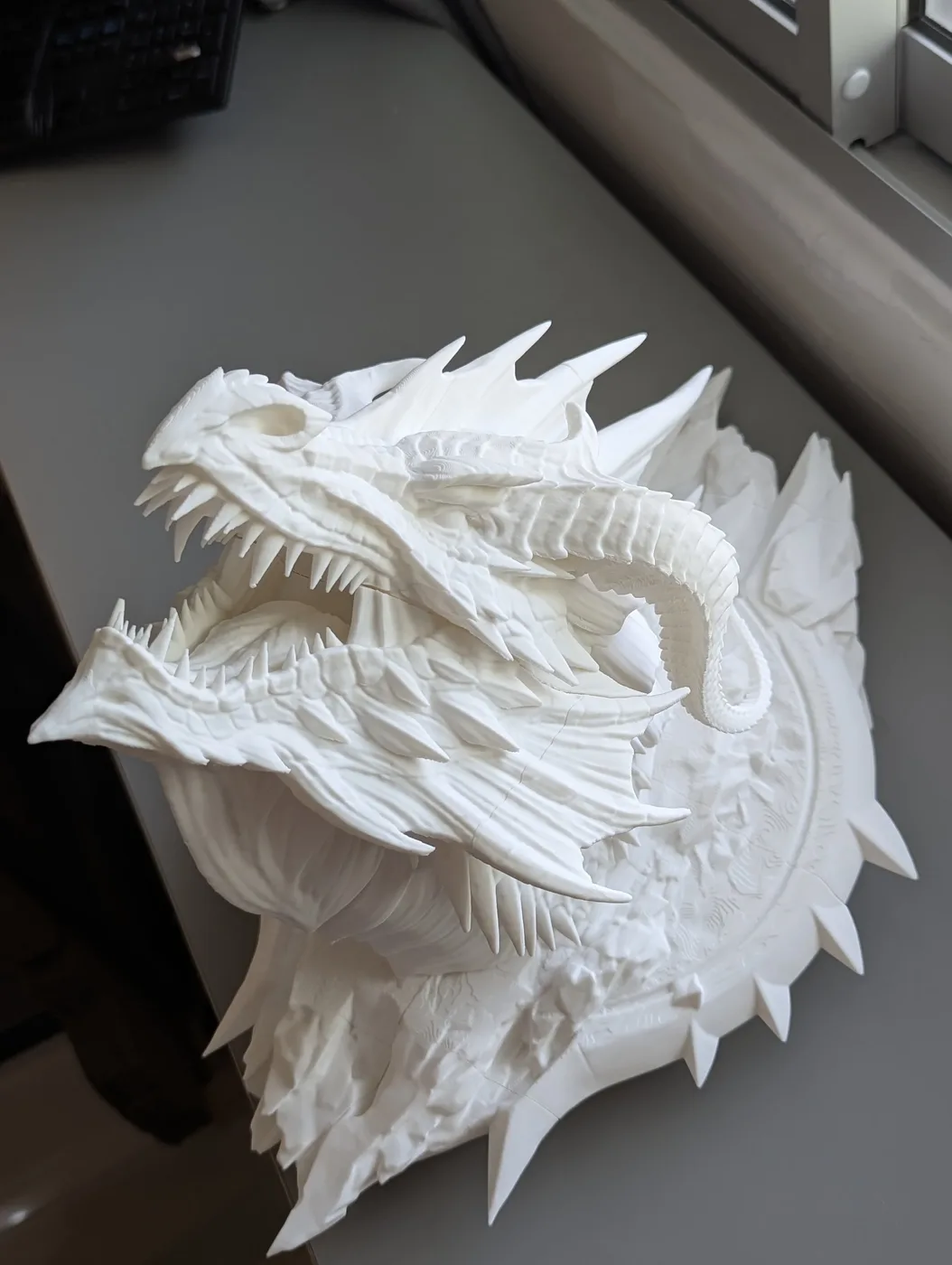 White 3D-printed sculpture of a dragon with detailed scales, teeth, and horns, showing an aggressive stance, on a desk near a window.